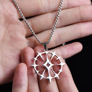 Puede incluir: Un collar con colgante plateado con un diseño circular con bordes puntiagudos y una forma de diamante central. El colgante cuelga de una cadena. La joya se sostiene en la mano de una persona.