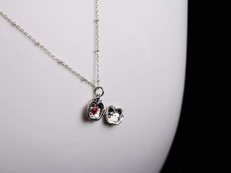 May include: A silver necklace with a pendant shaped like an apple. The apple pendant opens to reveal a red heart. The necklace is on a white background.