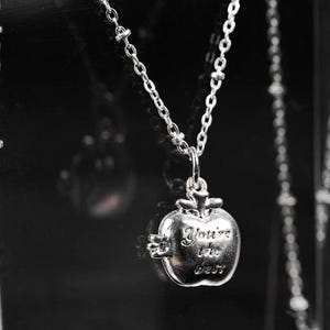May include: Silver-toned necklace featuring a pendant shaped like an apple. The apple pendant has the inscription "You're the best" and hangs from a delicate chain. The background is dark, highlighting the jewelry.