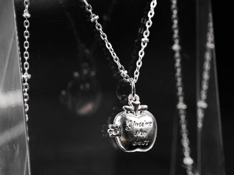 May include: Silver-toned necklace featuring a pendant shaped like an apple. The apple pendant has the inscription "You're the best" and hangs from a delicate chain. The background is dark, highlighting the jewelry.