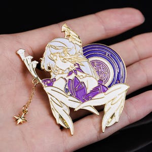 以下が含まれることがあります： 金色のエナメルピン。白い髪と紫色のアクセントを持つ、様式化されたアニメキャラクターが特徴です。ピンには星のチャームが付いたゴールドチェーンが付いています。デザインには幾何学模様の円形要素が含まれています。