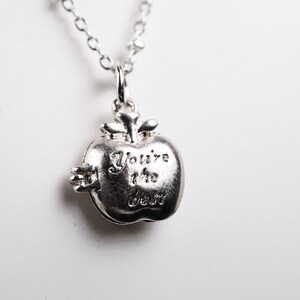 May include: Silver-toned apple pendant necklace with the words "You're the best" engraved on the front. The necklace has a delicate chain and a detailed apple charm, perfect for gifting.