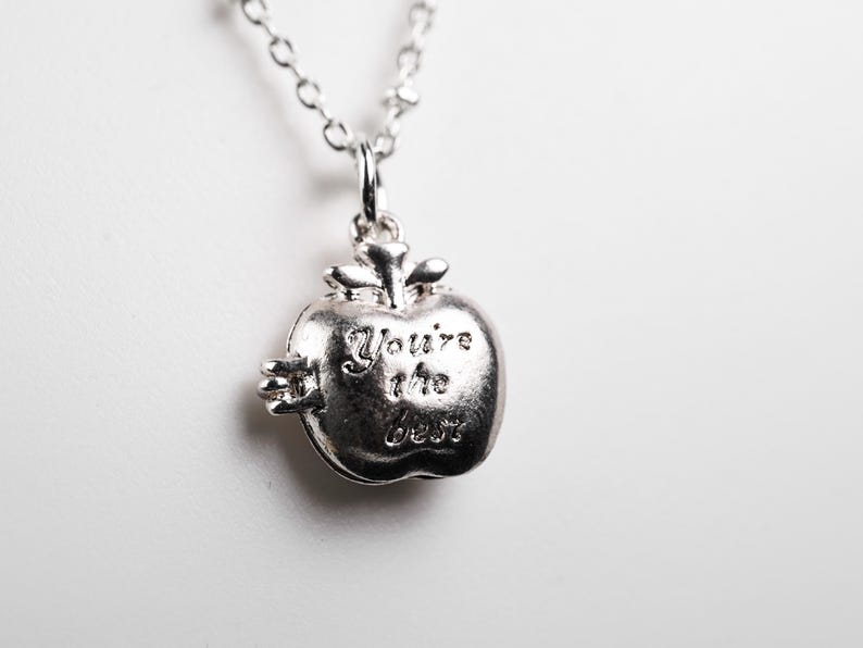 May include: Silver-toned apple pendant necklace with the words "You're the best" engraved on the front. The necklace has a delicate chain and a detailed apple charm, perfect for gifting.