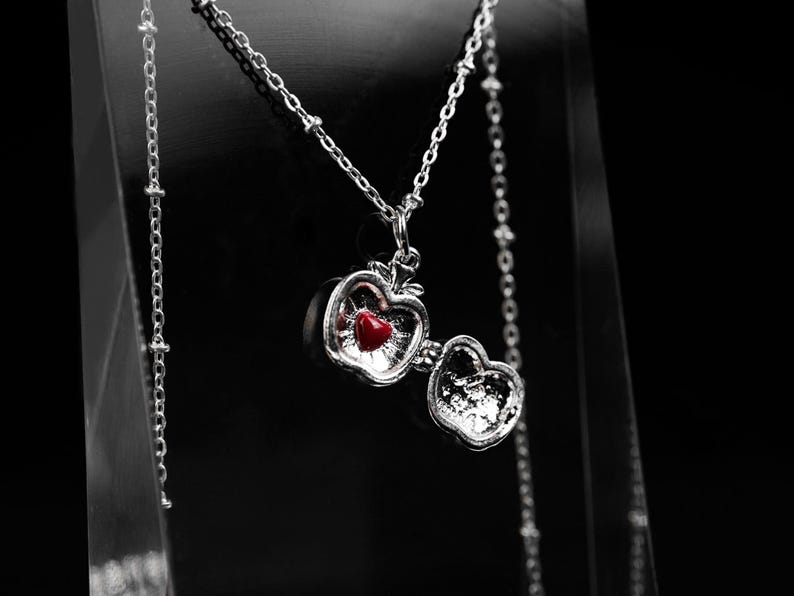 May include: A silver-toned necklace with a pendant shaped like an apple. The apple pendant opens to reveal a red heart. The necklace is displayed against a black background.