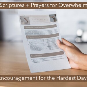 May include: A printed page titled "Scriptures + Prayers for Overwhelm" and "Encouragement for the Hardest Days" is held by a hand. The page contains text about finding peace and scriptures for the soul. The background is a blurred interior.