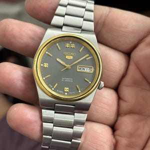 Może przedstawiać: Srebrny zegarek automatyczny Seiko 5 z szarą tarczą i złotymi akcentami. Zegarek wyświetla dzień i datę, ze srebrną bransoletą. Na tarczy zegarka widnieje napis "SEIKO 5 AUTOMATIC 21 JEWELS".