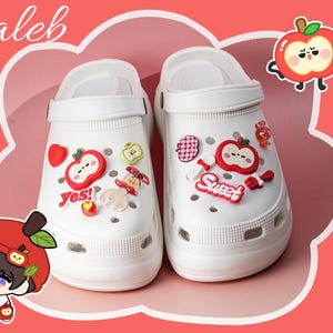 以下が含まれることがあります： リンゴをテーマにしたチャームと「Yes!」と「Sweet!」の文字で飾られた白いクロッグ。クロッグにはストラップが付いており、「Caleb」という名前が筆記体で書かれたピンク色の背景に配置されています。
