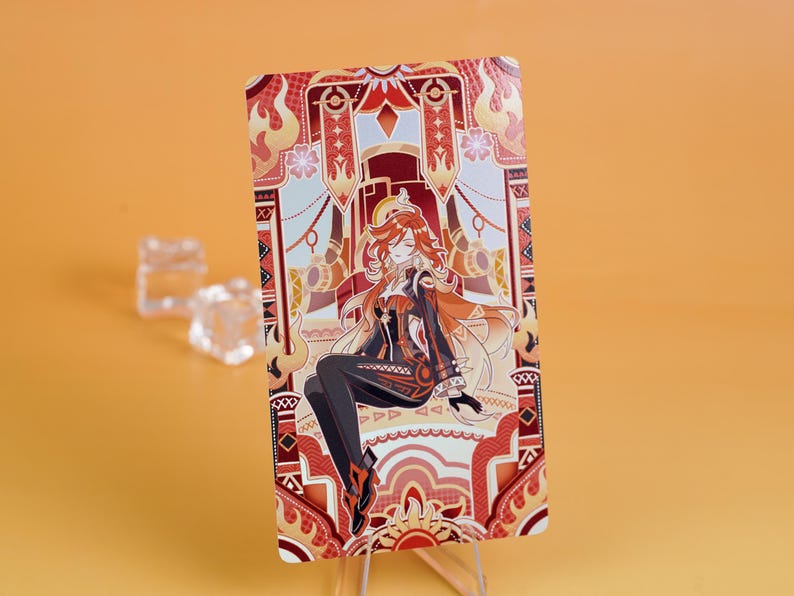 Puede incluir: Una tarjeta rectangular con una ilustraci&oacute;n estilizada de un personaje con cabello rojo y un atuendo negro. La tarjeta tiene un borde decorativo con tonos rojos, naranjas y dorados. El personaje est&aacute; sentado, y el fondo tiene patrones ornamentados.