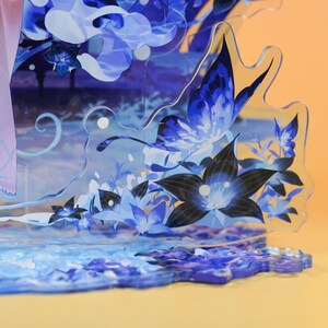 Op de afbeelding: Een gelaagd acryl kunstwerk met een vlinder en bloemenontwerp in blauw- en zwarttinten. Het kunstwerk heeft een waterachtige basis en een lichtoranje achtergrond. Het stuk is een decoratief item.