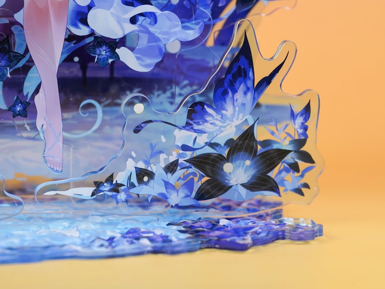 Op de afbeelding: Een gelaagd acryl kunstwerk met een vlinder en bloemenontwerp in blauw- en zwarttinten. Het kunstwerk heeft een waterachtige basis en een lichtoranje achtergrond. Het stuk is een decoratief item.