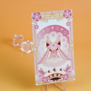 Puede incluir: Una tarjeta decorativa rosa y blanca con una ilustraci&oacute;n de un personaje con orejas largas y un vestido. La tarjeta tiene un dise&ntilde;o floral y celestial con detalles en rosa y dorado. Se muestra sobre un soporte transparente sobre un fondo naranja.