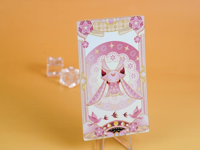 Puede incluir: Una tarjeta decorativa rosa y blanca con una ilustraci&oacute;n de un personaje con orejas largas y un vestido. La tarjeta tiene un dise&ntilde;o floral y celestial con detalles en rosa y dorado. Se muestra sobre un soporte transparente sobre un fondo naranja.