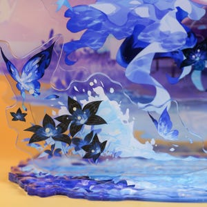 Op de afbeelding: Een decoratieve acryl display met een blauw-wit aquatisch thema. Het ontwerp bevat vlinders, bloemen en waterelementen. De basis heeft een golvend, gestructureerd ontwerp.