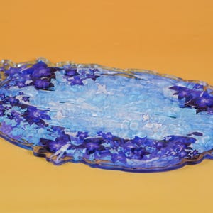 Op de afbeelding: Een decoratieve ovale schaal met een bloemenontwerp. De schaal heeft een lichtblauwe en witte achtergrond met donkerblauwe bloemaccenten langs de randen. De schaal is gemaakt van een transparant materiaal en ligt op een oranje oppervlak.