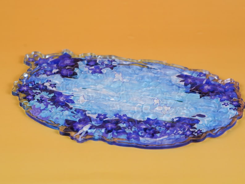 Op de afbeelding: Een decoratieve ovale schaal met een bloemenontwerp. De schaal heeft een lichtblauwe en witte achtergrond met donkerblauwe bloemaccenten langs de randen. De schaal is gemaakt van een transparant materiaal en ligt op een oranje oppervlak.