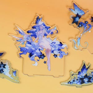Op de afbeelding: Een verzameling acrylfiguren met een fantasiekarakter in blauwe en witte kleding, omringd door blauwe vlinders en bloemmotieven. De figuren staan op een gele achtergrond.