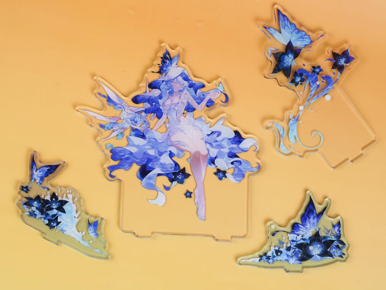 Op de afbeelding: Een verzameling acrylfiguren met een fantasiekarakter in blauwe en witte kleding, omringd door blauwe vlinders en bloemmotieven. De figuren staan op een gele achtergrond.