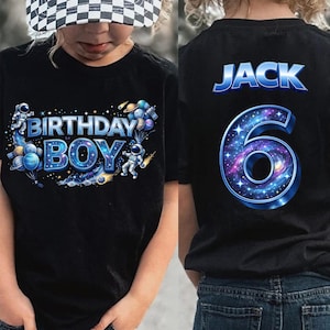 May include: Black t-shirt with a space-themed "BIRTHDAY BOY" graphic on the front and "JACK 6" on the back. The design includes astronauts, planets, and a galaxy-filled number 6. The child is wearing a black and white checkered baseball cap.