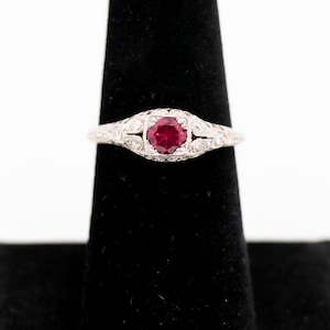 May include: A silver ring featuring a central, round, bright red gemstone, with smaller clear stones either side. The ring is displayed on a black velvet ring holder against a white backdrop. The ring has detailed, ornate design features.