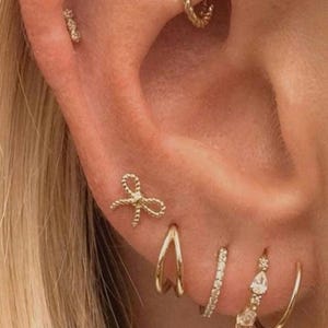 May include: Close-up of an ear displaying various gold earrings. The collection includes a bow stud, a textured stud, a small hoop, a V-shaped earring, a hoop with small stones, and a teardrop-shaped earring, showcasing a layered look.
