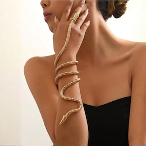 Puede incluir: Juego de brazalete y anillo de serpiente dorados. La joyería presenta un diseño detallado de serpiente que se envuelve alrededor del brazo y el dedo. La cabeza de la serpiente forma el anillo y el cuerpo se enrolla alrededor del brazo.