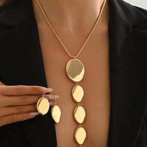 May include: Gold-toned jewelry set featuring a long necklace with four oval pendants and matching oval drop earrings. The necklace and earrings have a polished, reflective surface. The jewelry is displayed against a black background.