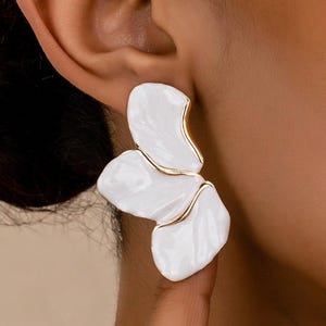 May include: White and gold-tone floral earrings. The earrings feature a three-petal design with a glossy white finish and gold-tone trim. The earrings are a statement piece, suitable for adding a touch of elegance to any outfit.