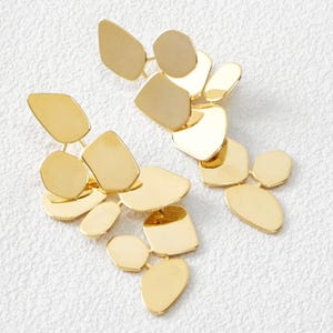 Puede incluir: Un par de pendientes dorados con un diseño abstracto. Cada pendiente presenta múltiples formas planas e irregulares conectadas por finas barras doradas. Los pendientes se muestran sobre una superficie blanca texturizada.