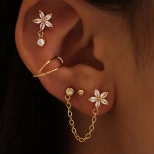 May include: An ear is adorned with a selection of gold-coloured earrings. The set features a flower-shaped stud with clear crystals, a dangling earring with a single crystal, a double-hoop ear cuff, and a chain connecting two earrings. The earrings are set against a warm skin tone.