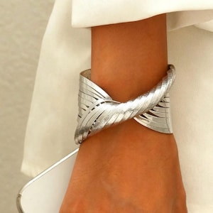 Può includere: Bracciale a polsino argentato con un design strutturato e stratificato. Il bracciale si avvolge attorno al polso, con un elemento centrale attorcigliato e molteplici bande parallele. Il gioiello è indossato su uno sfondo bianco.