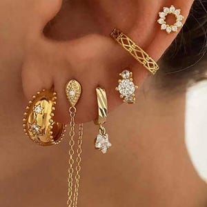 May include: A selection of gold-coloured earrings, including hoop earrings with star details, a chain earring, and a star-shaped dangle earring. Other earrings include a diamond-like stud and a diamond-like ring earring. The earrings are worn on the ear.
