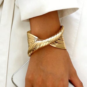 Könnte beinhalten: Ein goldfarbenes Armreif-Armband mit einem strukturierten, geschichteten Design. Das Armband umschließt das Handgelenk und weist ein zentrales, verdrehtes Element auf. Der Schmuck wird am Arm einer Person vor weißem Hintergrund getragen.