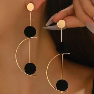 Può includere: Orecchini color oro dal design moderno. Ogni orecchino presenta un piccolo borchia rotonda, un accento circolare nero e una forma a semicerchio dorata. Gli orecchini sono mostrati su una persona.