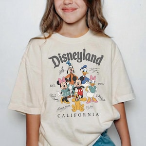 May include: Cream-colored Disneyland t-shirt featuring a graphic of Mickey Mouse, Minnie Mouse, Donald Duck, Daisy Duck, Goofy, and Pluto. The shirt has the text "Disneyland California" in a vintage font.