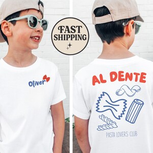 May include: White t-shirt with the name "Oliver" and a butterfly graphic on the front. The back of the shirt features the words "AL DENTE" and pasta illustrations, with "PASTA LOVERS CLUB" below. The shirt is worn by a child.
