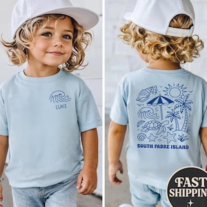 Op de afbeelding: Lichtblauw T-shirt met een golfpatroon en de naam "LUKE" op de voorkant. De achterkant toont een blauwe illustratie van een strandsc&egrave;ne met een schildpad, palmbomen en de tekst "SOUTH PADRE ISLAND".