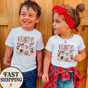 Puede incluir: Dos niños con camisetas blancas con un gráfico colorido con temática navideña con personajes de dibujos animados. El gráfico incluye la palabra "Merry" y varios personajes. Los niños sonríen y llevan vaqueros azules. Una pegatina de "Fast Shipping" es visible.
