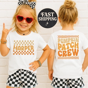 Pode incluir: Duas crianças vestindo camisetas brancas com os textos "HARPER" e "PUMPKIN PATCH CREW" em designs xadrez laranja e bege. Elas também estão usando shorts xadrez e acessórios.