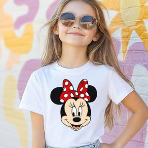Może przedstawiać: Biała koszulka z grafiką Myszki Minnie. Grafika przedstawia twarz Myszki Minnie z czerwoną kokardą w białe grochy. Osoba na zdjęciu ma na sobie okulary przeciwsłoneczne i stoi przed kolorową ścianą.