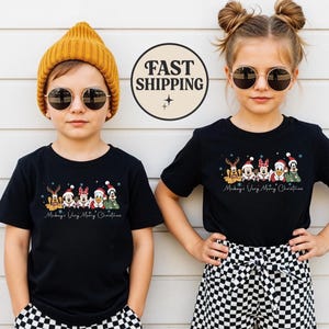 Puede incluir: Dos niños con camisetas negras con un diseño navideño. Las camisetas muestran personajes de Disney con gorros de Papá Noel y el texto "Mickey's Very Merry Christmas". Uno lleva un gorro amarillo, el otro tiene el pelo recogido en moños. Ambos llevan gafas de sol.