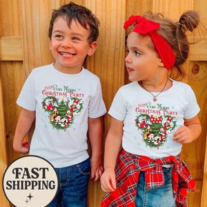Puede incluir: Dos niños con camisetas blancas con un diseño navideño de Mickey Mouse y sus amigos. Las camisetas dicen "Mickey's Very Merry Christmas Party". Los niños sonríen y llevan vaqueros azules. Una camisa de cuadros roja está atada a la cintura de un niño.