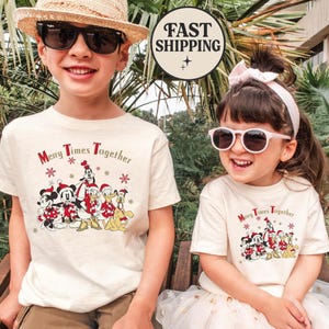 Puede incluir: Dos niños con camisetas color crema con un gráfico de personajes de Disney con gorros de Papá Noel. Las camisetas dicen "Merry Times Together" y presentan a Mickey Mouse, Minnie Mouse, Donald Duck y Goofy. Un niño lleva gafas de sol y un sombrero de paja.