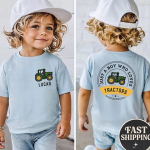 May include: A light blue t-shirt featuring a green and yellow tractor graphic. The front of the shirt has the name "LUCAS" below the tractor. The back of the shirt has a circular design with the text "JUST A BOY WHO LOVES TRACTORS".