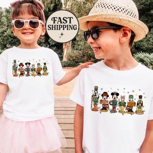 May include: Two children wearing white t-shirts with a colorful graphic of Disney characters dressed as nutcrackers. The graphic includes Mickey Mouse, Donald Duck, and Goofy. One child wears a pink tutu and sunglasses. A "FAST SHIPPING" logo is visible.