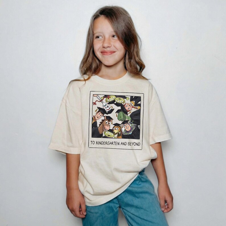 May include: A cream-colored short-sleeve t-shirt featuring a square graphic print of various animated characters wearing black graduation caps. Below the illustration, the text reads TO KINDERGARTEN AND BEYOND in a simple, bold, black sans-serif font.
