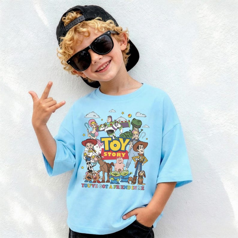 Op de afbeelding: Een lichtblauw T-shirt met een kleurrijke afbeelding van personages uit de film "Toy Story". Het shirt heeft korte mouwen en de tekst "Toy Story" en "You've Got a Friend in Me". Het shirt wordt gedragen door een persoon met een zwarte pet en zonnebril.