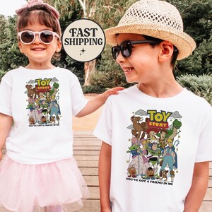 Puede incluir: Dos ni&ntilde;os con camisetas blancas con un gr&aacute;fico colorido de "Toy Story". El gr&aacute;fico incluye varios personajes de la pel&iacute;cula y el texto "YOU'VE GOT A FRIEND IN ME". Un ni&ntilde;o lleva un tut&uacute; rosa y gafas de sol, el otro lleva un sombrero de paja y gafas de sol.