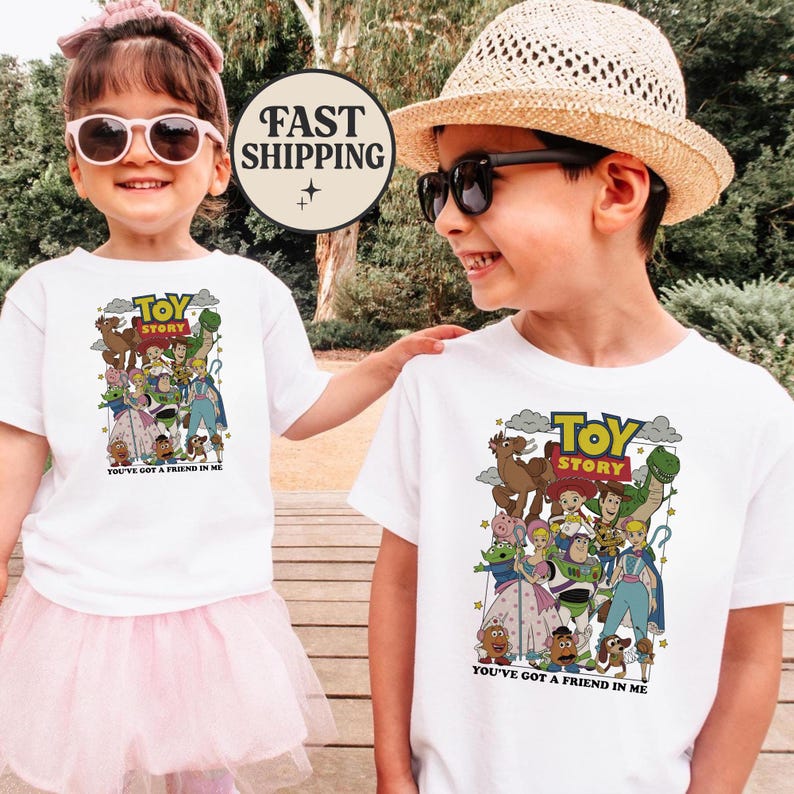 Puede incluir: Dos ni&ntilde;os con camisetas blancas con un gr&aacute;fico colorido de "Toy Story". El gr&aacute;fico incluye varios personajes de la pel&iacute;cula y el texto "YOU'VE GOT A FRIEND IN ME". Un ni&ntilde;o lleva un tut&uacute; rosa y gafas de sol, el otro lleva un sombrero de paja y gafas de sol.