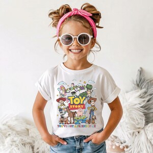 Op de afbeelding: Een wit T-shirt met een kleurrijke afbeelding van personages uit de animatiefilm "Toy Story". Het shirt heeft de woorden "Toy Story" in grote gele en blauwe letters, met de zin "You've Got a Friend in Me" eronder.