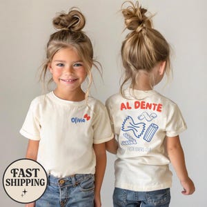 May include: Two children wearing cream-colored t-shirts with blue jeans. The front of the shirt has the name "Olivia" and a butterfly graphic. The back of the shirt has the words "AL DENTE" and pasta illustrations, with "PASTA LOVERS CLUB" below.